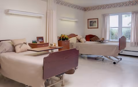A bright and clean patient room with two hospital beds, each with beige bedding and pillows. There is a wooden bedside table between the beds with a flower arrangement and a framed photo. The room has large windows with curtains, a framed picture on the wall, and a privacy curtain that can be drawn between the beds.