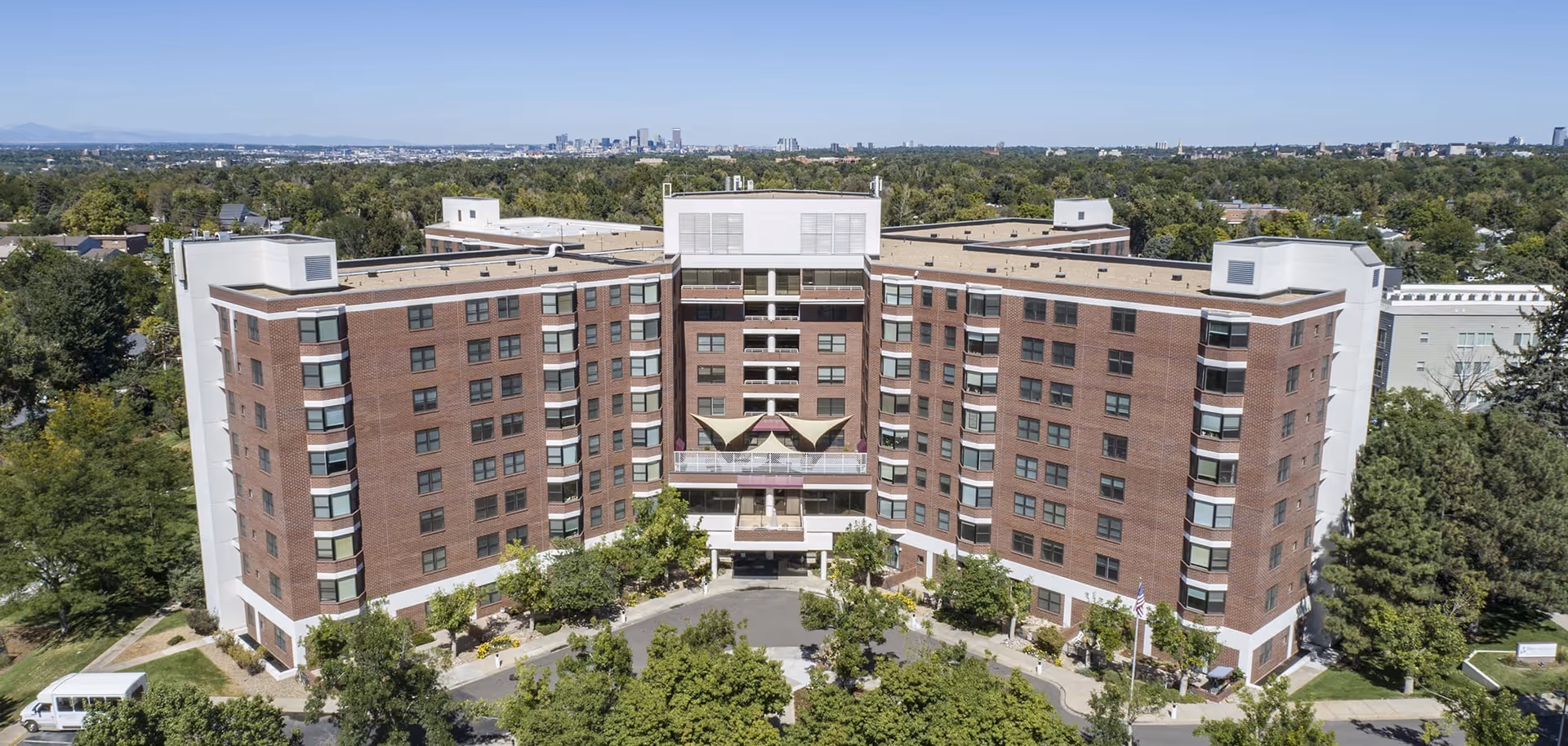 Aerial view of a large multi-story brick building with a U-shaped design surrounded by trees and greenery. The building has multiple windows and a central entrance with a covered area. In the background, a city skyline is visible under a clear blue sky.