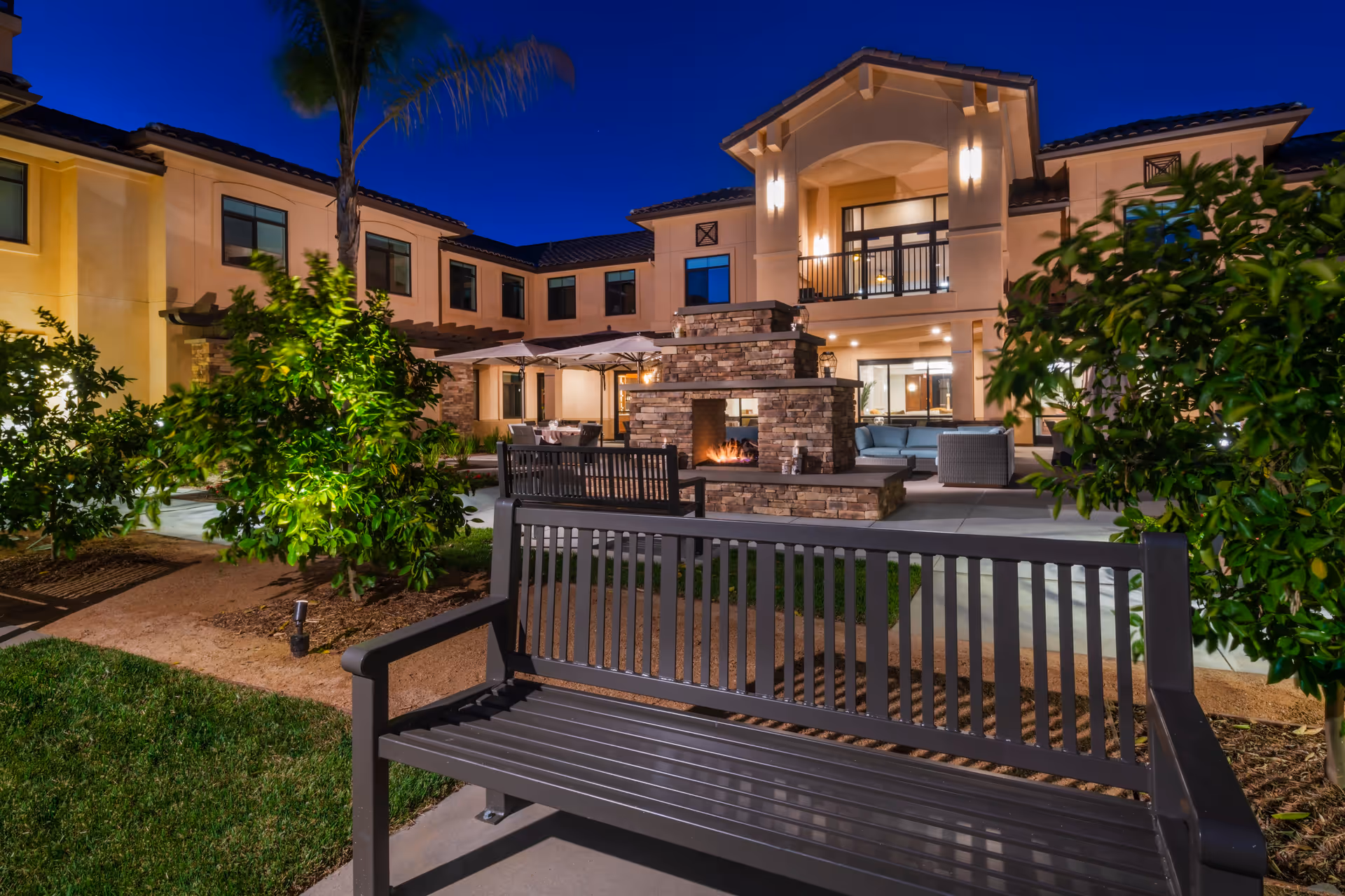 Night view of a courtyard at a senior living facility with benches, a stone fireplace, outdoor seating, and a lit two-story building facade.
