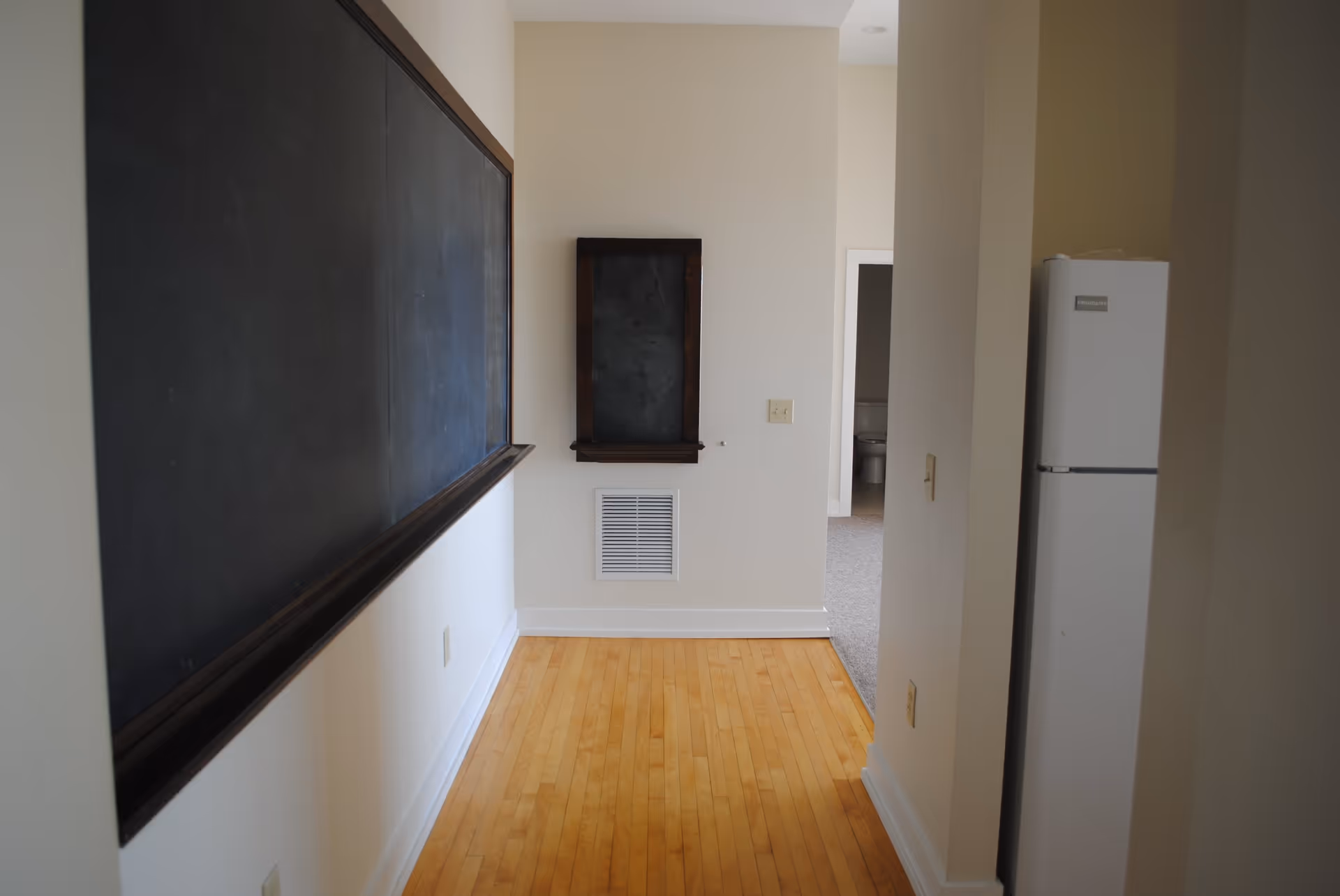 A narrow hallway with light wooden flooring and cream-colored walls. On the left wall, there is a large blackboard with a wooden frame. At the end of the hallway, there is a small blackboard or chalkboard mounted on the wall above a vent. To the right, a white refrigerator is partially visible, and an open doorway reveals a room with carpeted flooring and a toilet.
