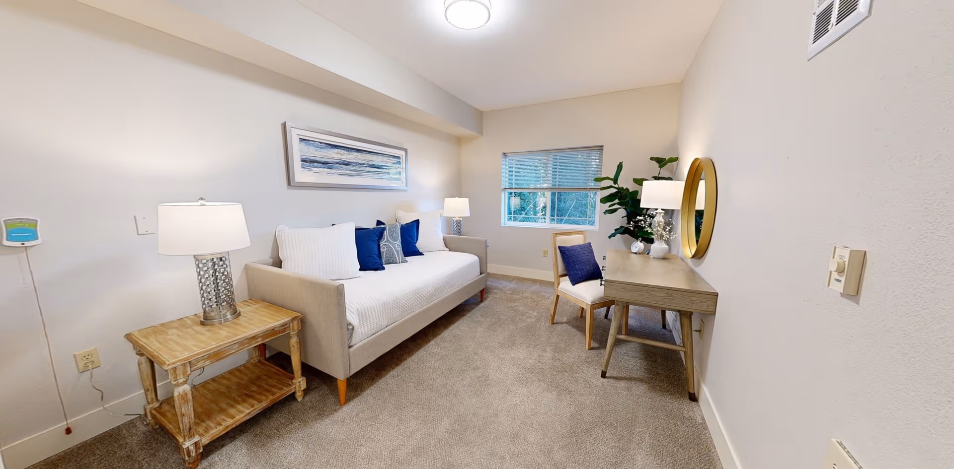 A cozy living room area with a beige sofa adorned with white and blue pillows, a wooden side table with a lamp, a wooden desk with a chair, a round mirror on the wall, and a window showing greenery outside. The room has beige carpet and light-colored walls.