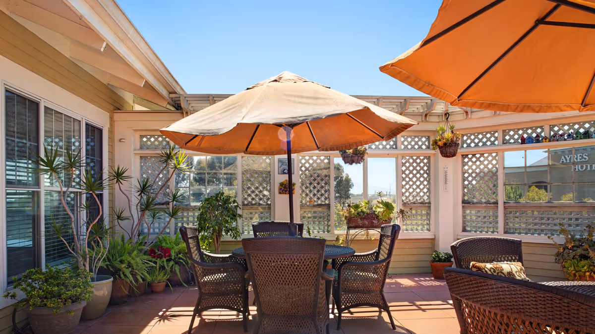 Outdoor patio area with a round table and six wicker chairs under a large beige umbrella. The patio is surrounded by lattice fencing with potted plants and hanging planters. The sky is clear and blue.