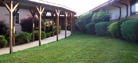 A covered walkway with wooden posts leading to the entrance of a building, surrounded by green grass and neatly trimmed bushes on either side.