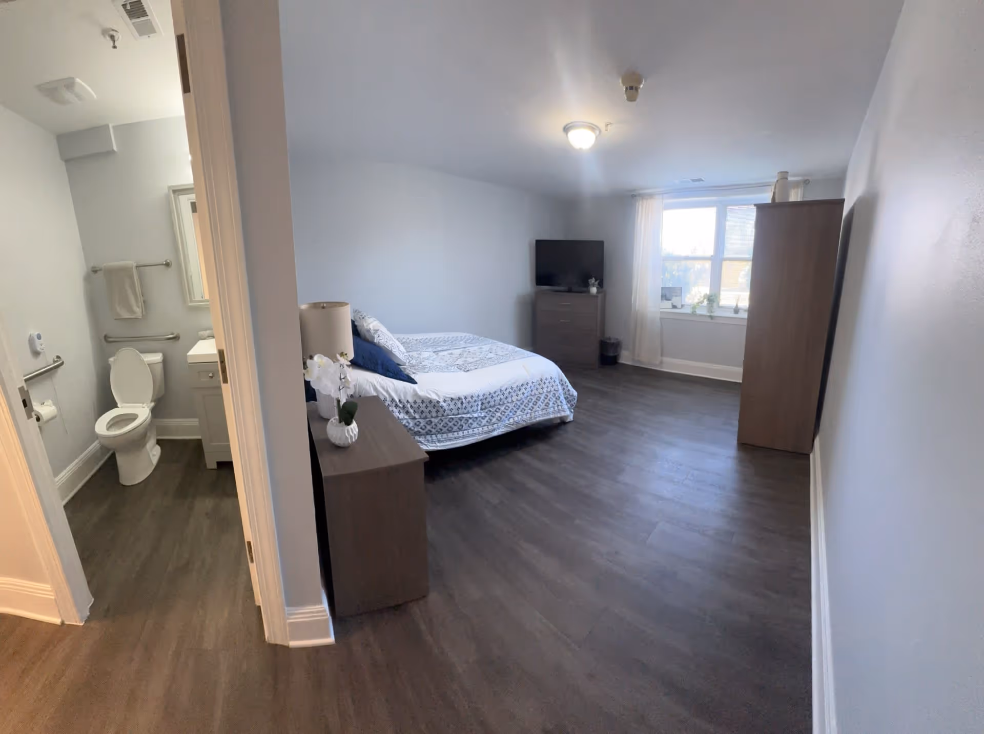 A senior living community room featuring a bedroom with a bed, nightstand with a lamp and flower, dresser with a TV, and a window with sheer curtains. To the left, there is an open door leading to a bathroom with a toilet, towel rack, and sink.