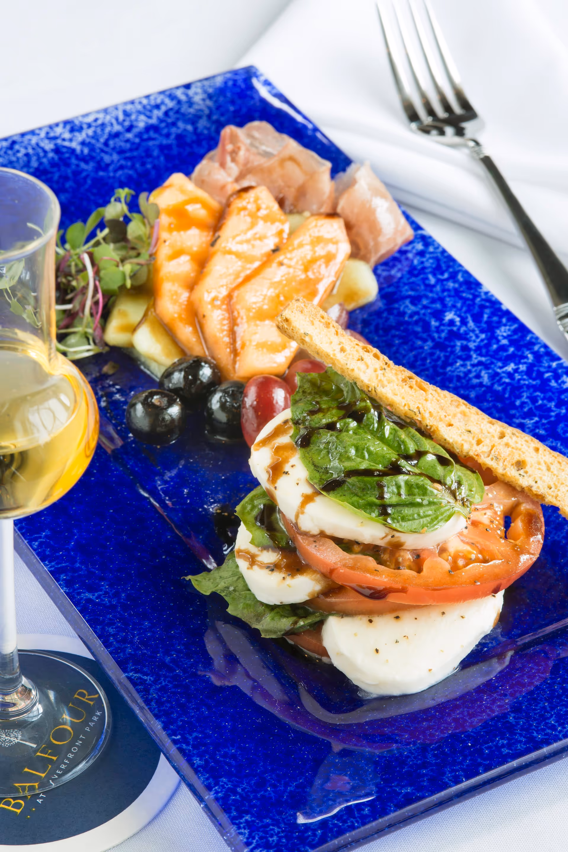 A gourmet plate featuring a caprese salad with fresh mozzarella, tomato slices, basil leaves, and a drizzle of balsamic glaze, accompanied by grapes, black olives, and slices of smoked salmon on a vibrant blue rectangular plate. A glass of white wine and a fork on a white napkin are visible in the background.