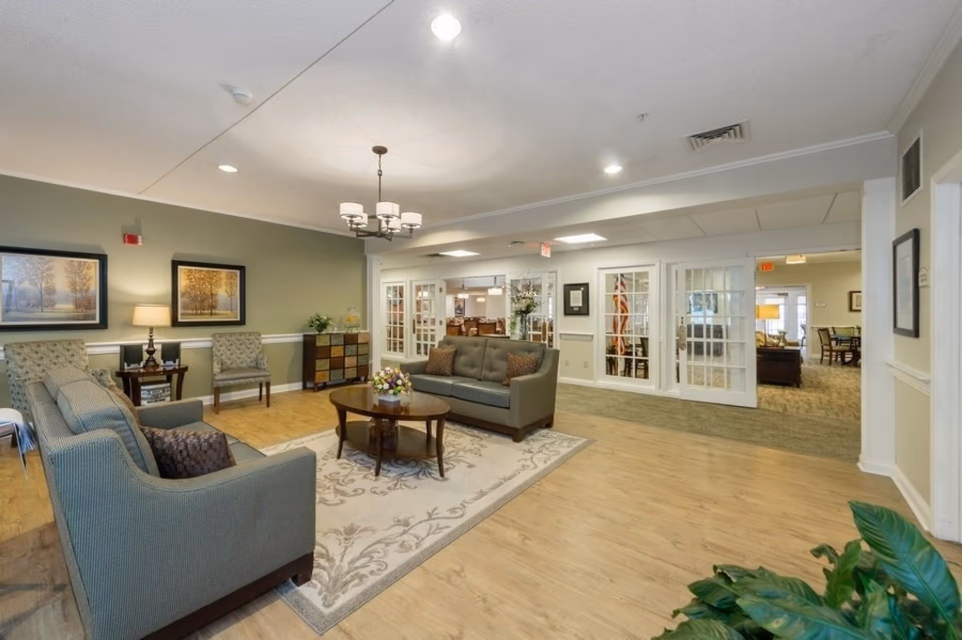 A spacious and well-lit living room area in Sycamore Manor featuring two gray sofas with decorative pillows, a wooden coffee table with a floral centerpiece on a patterned area rug, two armchairs, framed artwork on a green accent wall, a side table with a lamp, and a multi-colored drawer cabinet. The room has light wood flooring and white trim, with glass-paneled doors leading to other rooms in the background.