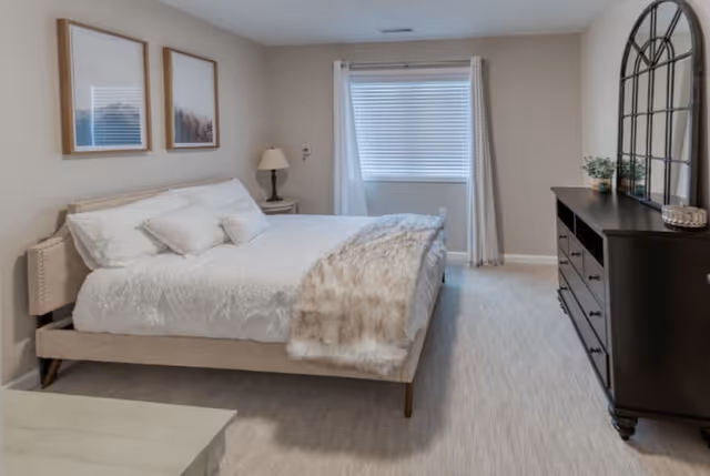 A neatly arranged bedroom with a large bed covered in white bedding and a light brown fur throw. There are two framed pictures on the beige wall above the bed. A bedside table with a lamp is next to the bed. Across from the bed is a dark wooden dresser with a large decorative mirror and small plants on top. A window with white curtains and blinds is centered on the far wall.