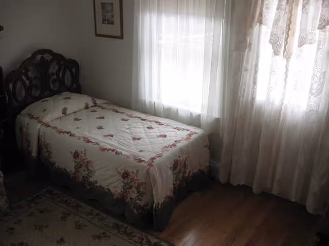 A small bedroom with a single bed covered in a floral patterned quilt. The bed has a dark wooden headboard with intricate designs. There is a window with sheer white curtains allowing natural light to enter the room. A framed picture hangs on the wall above the bed, and a floral rug is partially visible on the wooden floor.