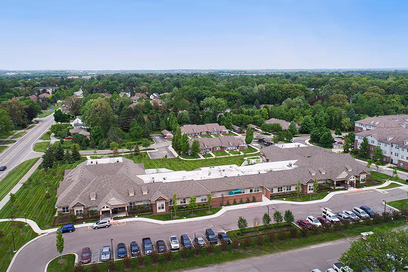 Aerial view of American House Freedom Place Rochester senior living facility showing a large single-story building with a parking lot in front, surrounded by green lawns, trees, and a suburban neighborhood in the background under a clear sky.