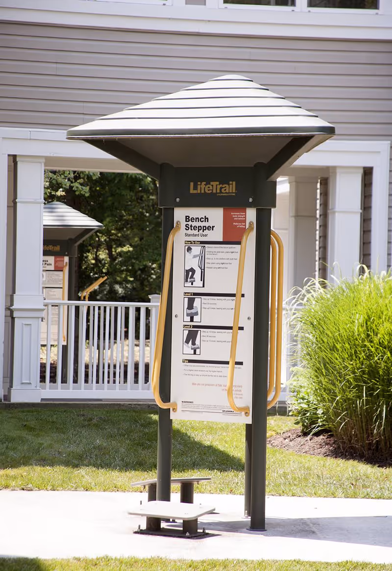 Outdoor exercise equipment labeled 'Bench Stepper' with instructions, situated on a concrete path with grass and bushes nearby, and a white railing and building in the background.