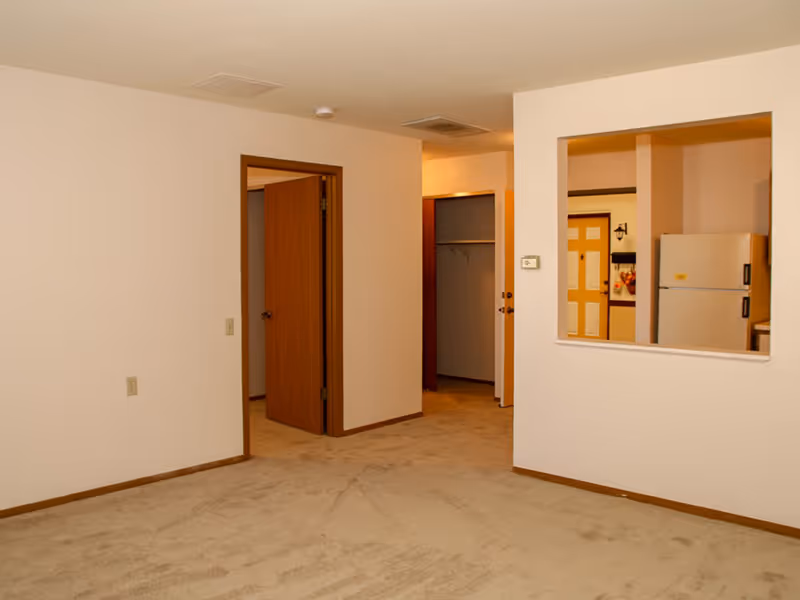 An empty room with beige carpet and off-white walls featuring a wooden door slightly ajar leading to another room. There is a rectangular pass-through window in the wall showing a white refrigerator and a door with a window in the background.