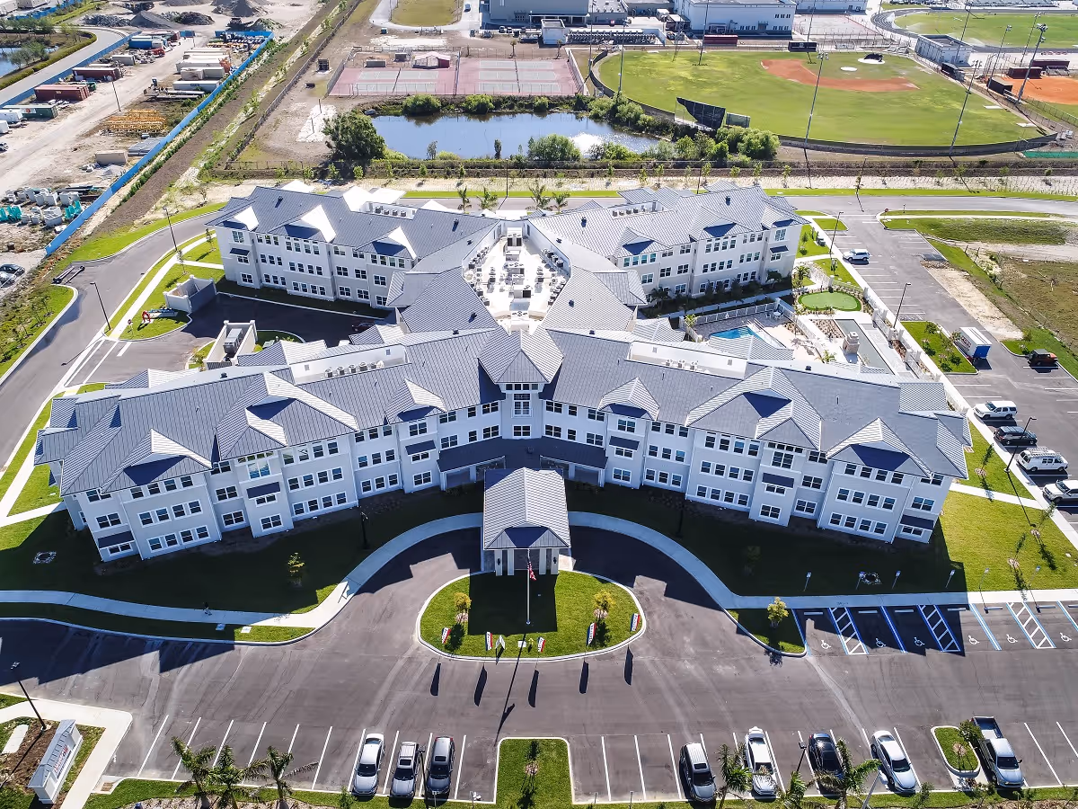 Aerial view of a large, modern senior living facility with multiple wings and a central courtyard. The building is surrounded by green lawns, parking lots with cars, and nearby sports fields including tennis courts and a baseball field. The facility has a gray roof and white exterior walls.