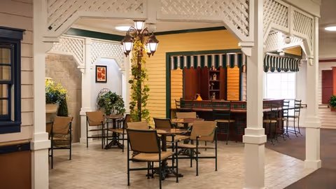 Indoor seating area designed to resemble an outdoor patio with tables and chairs, a lamppost with greenery, and a small bar or counter area with striped awnings and shelves in the background.