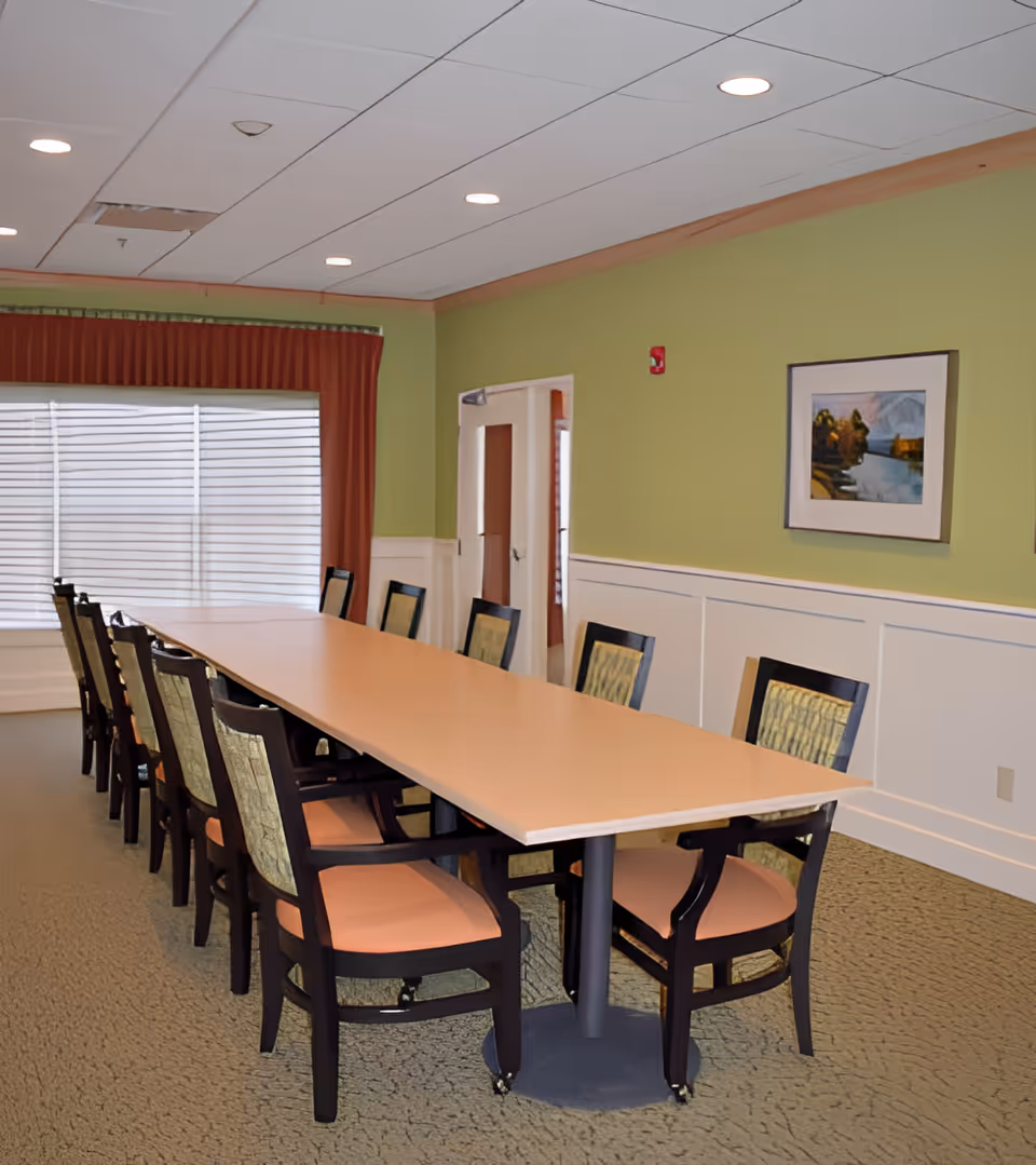 A long rectangular conference or dining table with twelve chairs arranged around it in a room with green walls, a large window with blinds and red curtains, a framed landscape painting on the wall, and a beige carpeted floor.