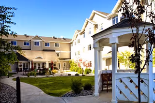 Outdoor courtyard area of a senior living facility with a paved walkway, green lawn, trees, shrubs, and a white gazebo. The multi-story building with beige siding and multiple windows surrounds the courtyard under a clear blue sky.