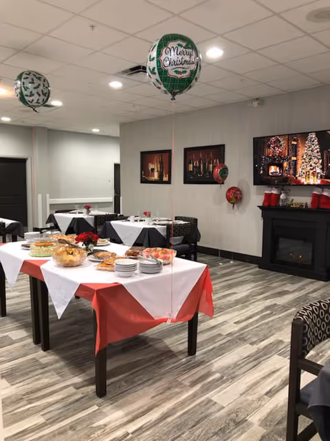 A holiday-decorated dining room with a buffet table, dining tables, balloons, and a fireplace with a TV showing a Christmas scene.