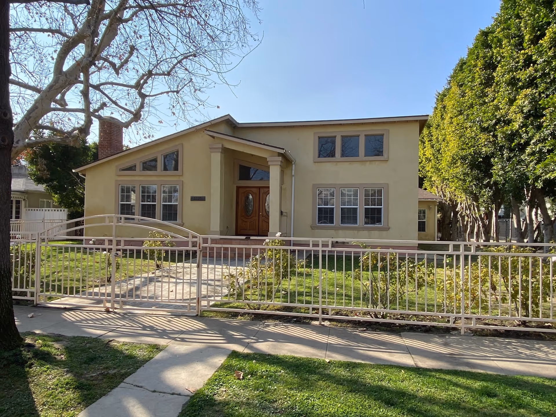 Front exterior view of a single-story beige building with a sloped roof, multiple windows, and a double wooden front door. The building is surrounded by a metal fence with a gate, a green lawn, some bushes, and trees. The sky is clear and blue.