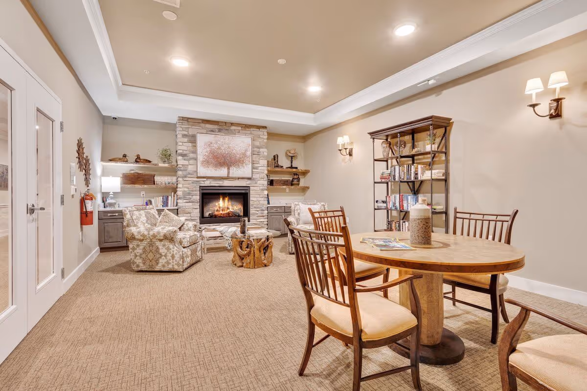 Cozy communal living room with a stone fireplace, armchairs, bookshelves, and a round wooden table with chairs.