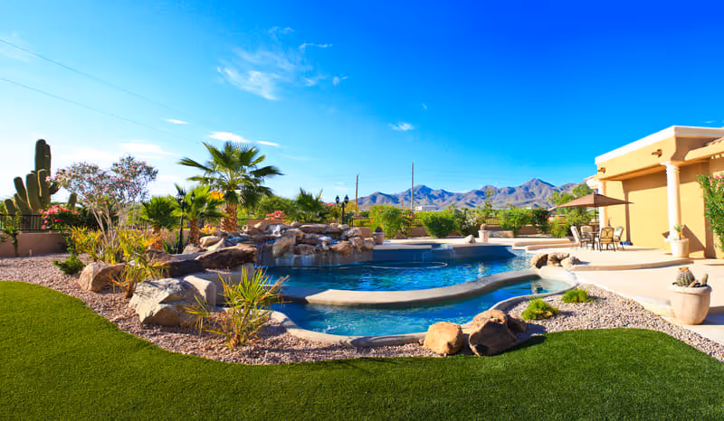 Outdoor view of a senior living facility featuring a swimming pool with a rock waterfall, surrounded by desert landscaping including cacti and palm trees, with mountains visible in the background under a clear blue sky.