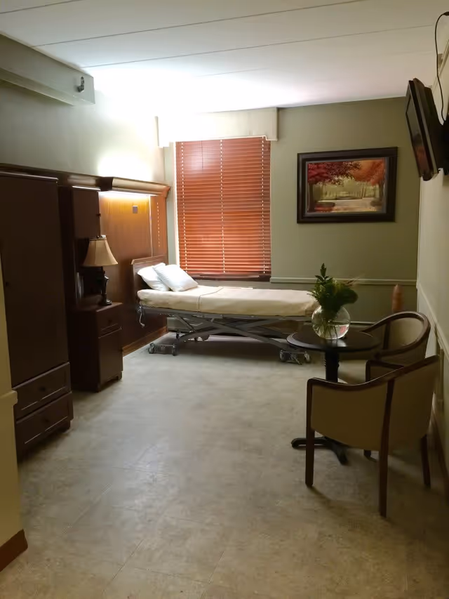 A small room with a medical bed positioned near a window with closed wooden blinds. The room has beige walls and tiled flooring. There is a wooden cabinet and a nightstand with a lamp on the left side. On the right side, there is a small round table with a vase of flowers and two chairs. A framed painting of a forest scene hangs on the wall, and a TV is mounted on the upper right corner.