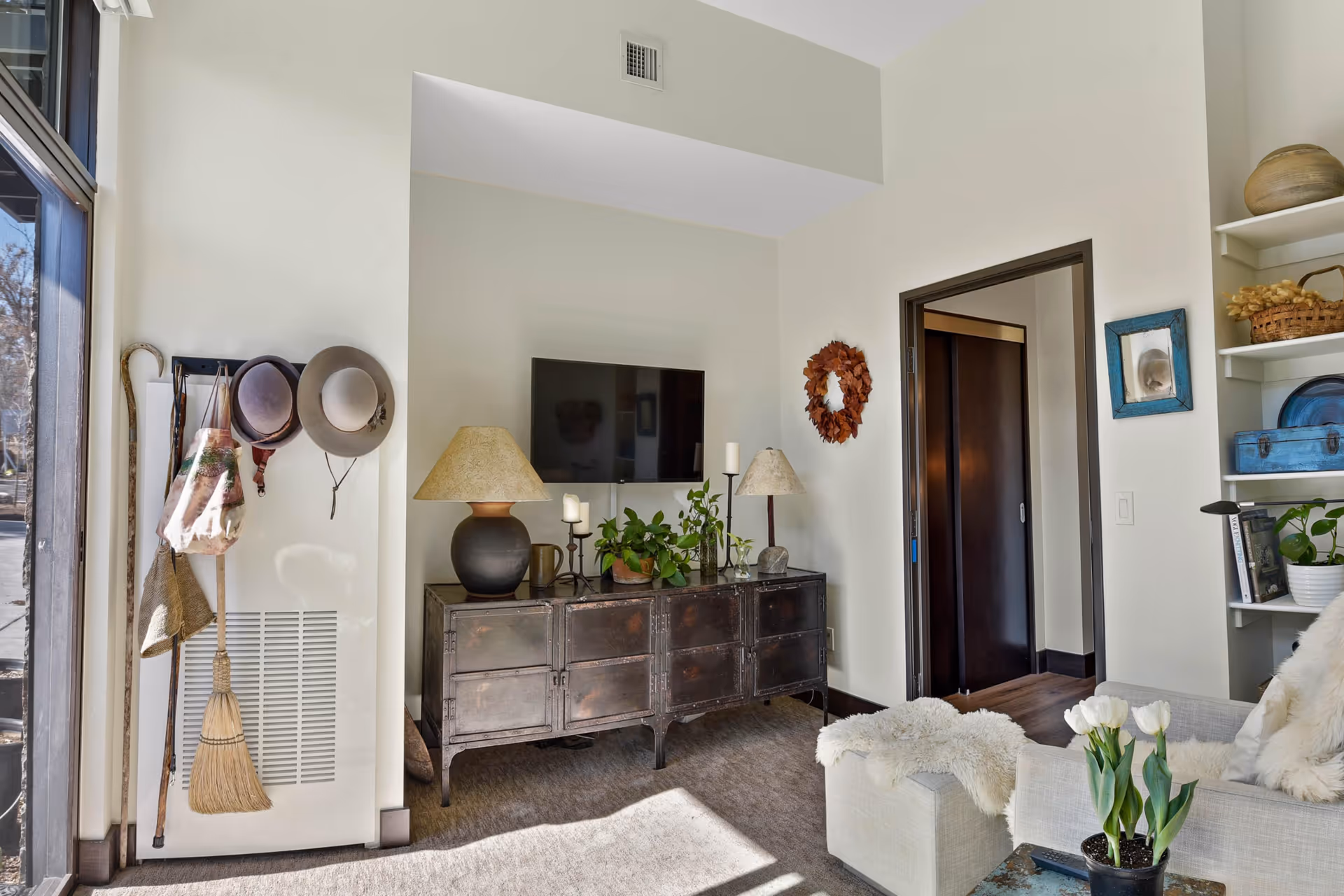 Bright living room with a mounted TV above a rustic cabinet, lamps and plants, hats and a broom hung by a large window, and a cozy sofa with a coffee table holding tulips.