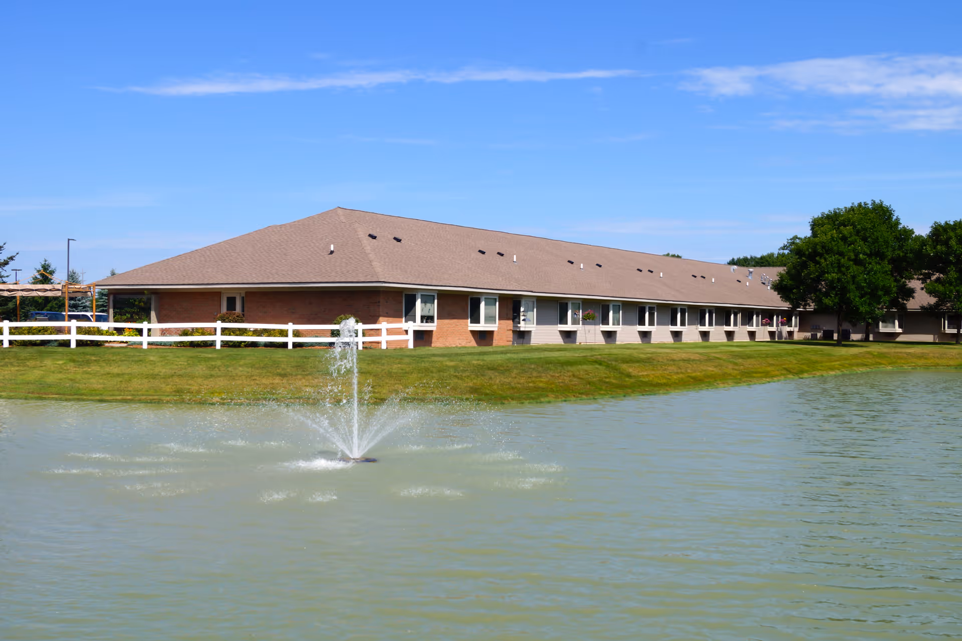 Long single-story assisted living building beside a pond with a fountain, grassy lawn, and white fence under a blue sky.