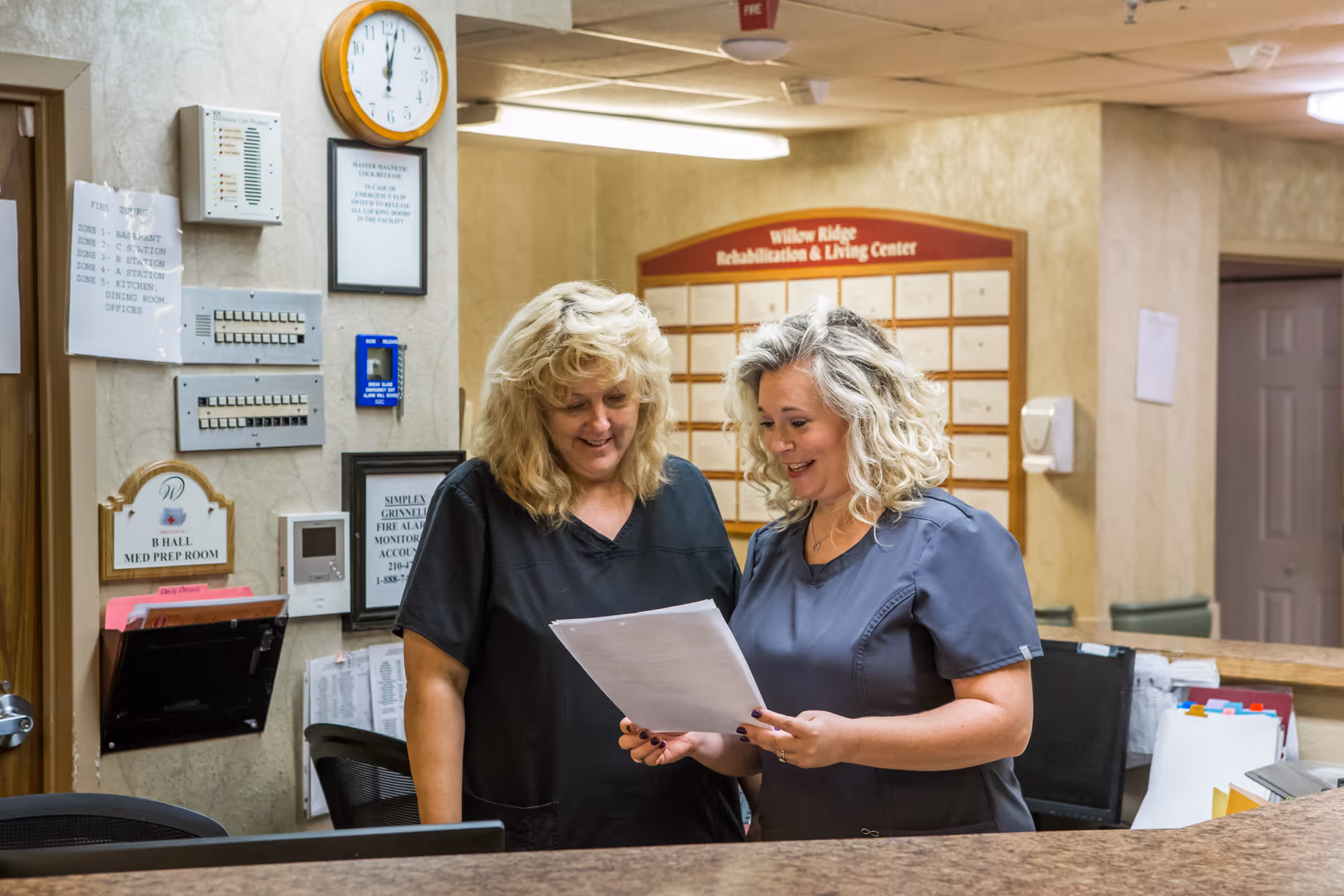 Two female healthcare workers in scrubs standing behind a reception desk, looking at and discussing a document. The background shows a wall with a clock, various notices, and a board labeled 'Willow Ridge Rehabilitation & Living Center.'