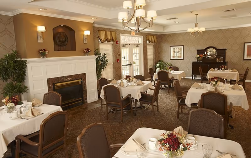 A cozy dining room with several tables covered in white tablecloths, each set with cups, napkins, and floral centerpieces. The room features a fireplace with decorative plants on either side, patterned wallpaper, and warm lighting from wall sconces and chandeliers. There are multiple chairs around each table and framed artwork on the walls.