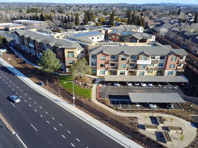 Aerial view of Prairie City Landing, a multi-story residential building with a parking lot featuring covered parking spaces. The building is surrounded by landscaped areas with trees and pathways, and a road runs alongside the property. The surrounding neighborhood includes many houses and trees extending into the distance.