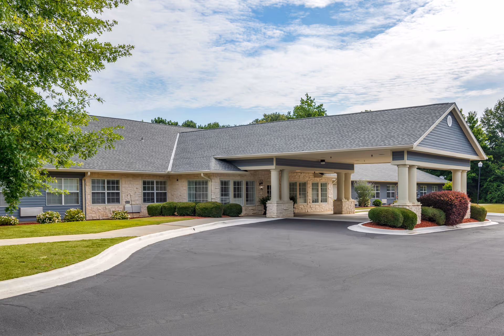 Front entrance of a single-story senior living building with a covered porte-cochere, landscaped shrubs, and a paved driveway.