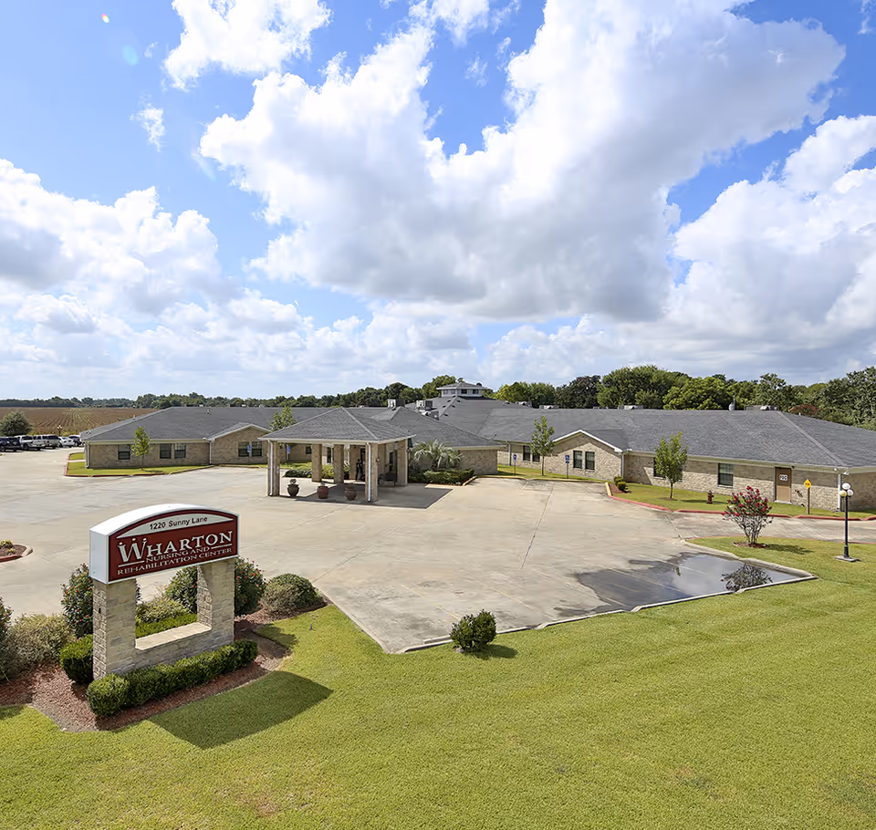 Exterior view of Wharton Nursing and Rehabilitation Center, a single-story building with a large parking area in front, surrounded by green grass and trees under a partly cloudy blue sky.