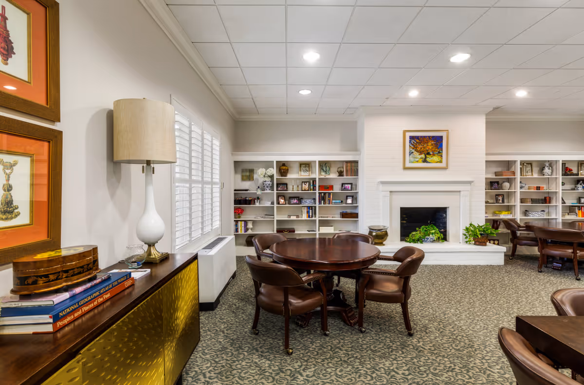 A cozy common area with a round wooden table surrounded by four brown leather chairs on a patterned carpet. The room features a white brick fireplace with a colorful painting above it, flanked by built-in white bookshelves filled with books, vases, and framed photos. To the left, a sideboard with a white lamp, decorative boxes, and stacked books sits beneath two framed artworks. The room is softly lit with recessed ceiling lights and has white plantation shutters on the window.