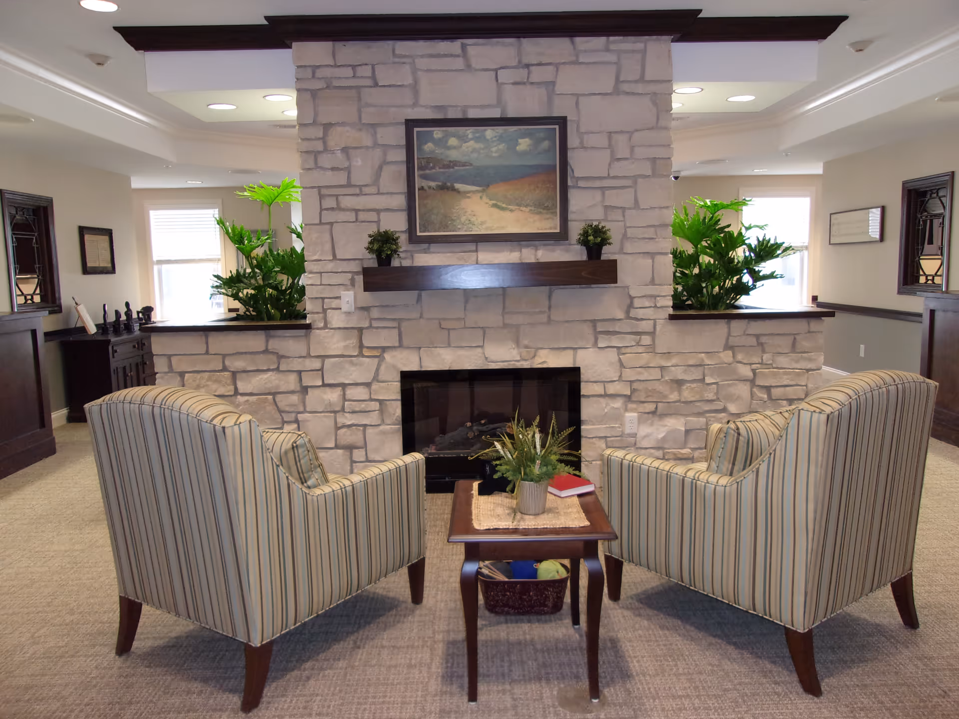 Two striped armchairs face a stone fireplace with a small table, plants, and framed art in a comfortable lounge.
