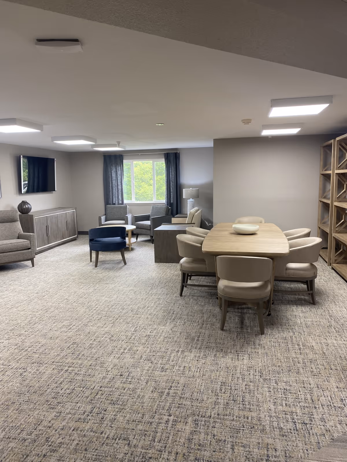 A spacious living room area with a dining table and six chairs on the right side, a seating area with armchairs and a small round table near a window with dark curtains, a wall-mounted TV, and a wooden cabinet beneath it. The room has neutral-colored walls and carpeted flooring.