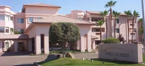 Front view of a pink senior living building with palm trees and entry drive.