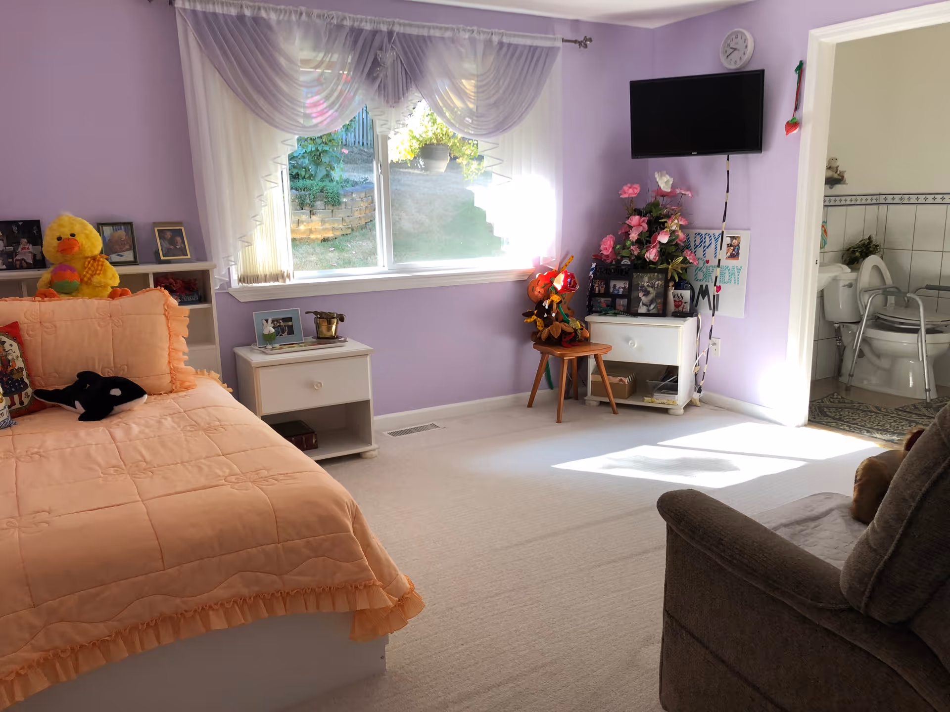 Sunlit lavender bedroom with a peach bedspread, nightstands, a wall-mounted TV, and an open doorway to an adjoining bathroom.
