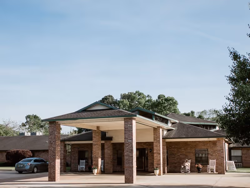 Front exterior view of Summit Nursing & Rehab of San Augustine showing a covered entrance supported by brick columns, a driveway with a parked car, and two rocking chairs near the entrance. Trees and a clear sky are visible in the background.