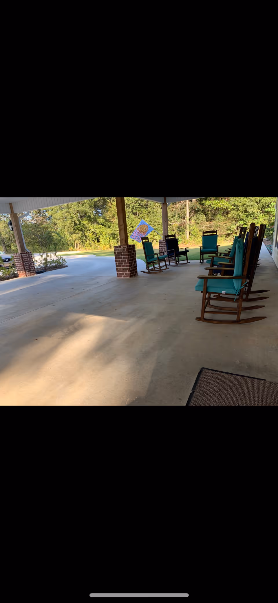 Covered outdoor patio area with several wooden rocking chairs with green cushions arranged along the edge. The patio has brick pillars and overlooks a green, wooded area with trees and grass.