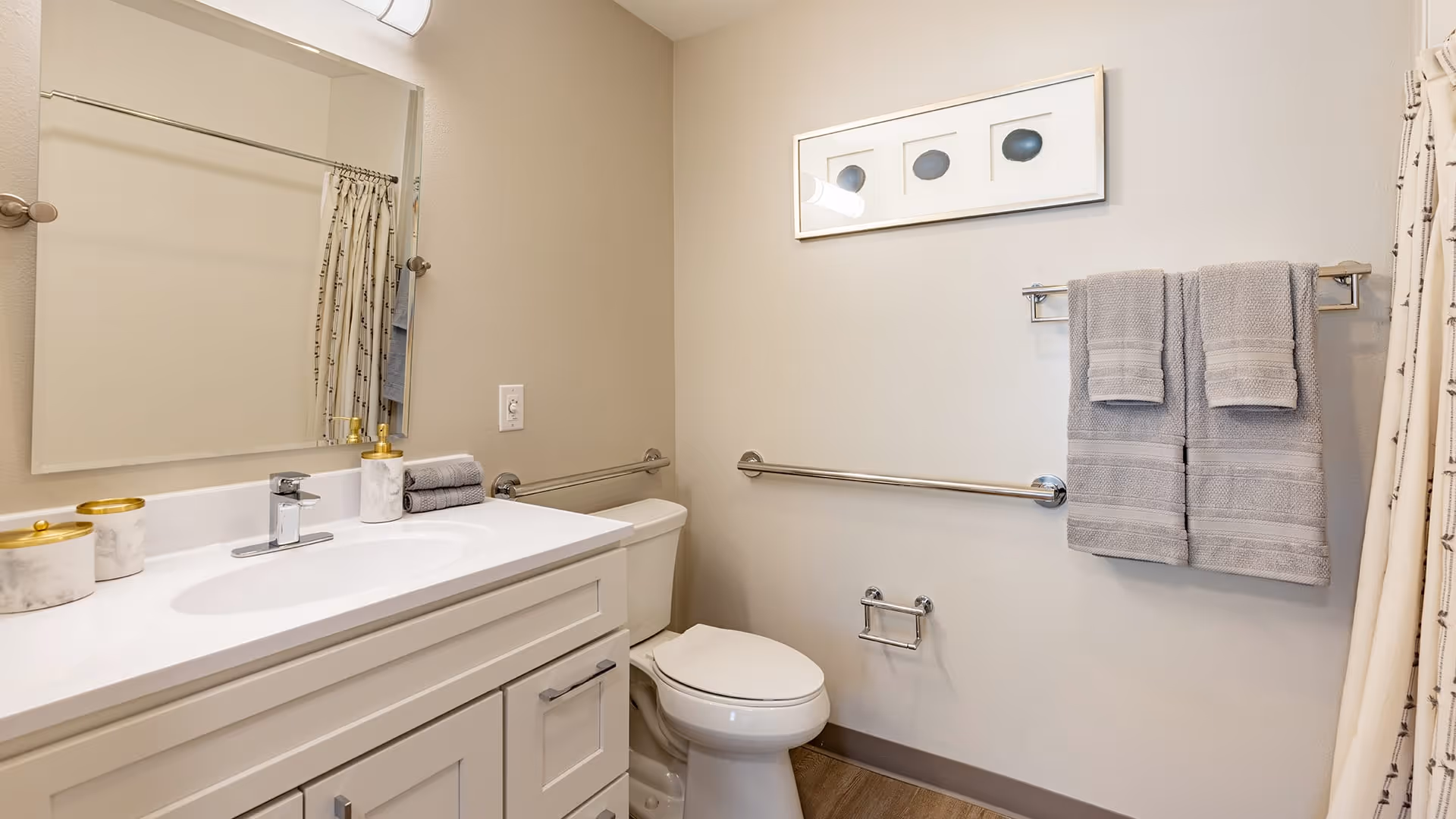 A clean and modern bathroom featuring a white vanity with a sink, a large mirror above it, a toilet, and a towel rack with two gray towels. The walls are light-colored, and there is a framed artwork above the towel rack. A shower curtain is partially visible on the right side.
