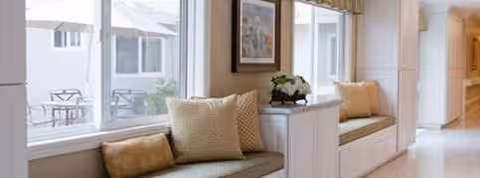 Sunlit window-seat lounge with cushions, framed art, and a potted plant along a hallway.