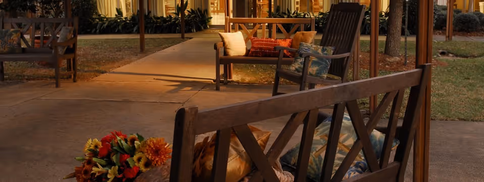 Outdoor seating area with wooden benches and chairs with cushions along a walkway leading to a lit building entrance.