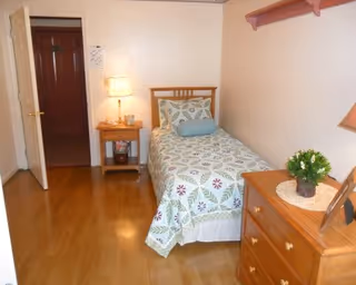 A small bedroom with a single bed covered in a patterned bedspread with a cylindrical pillow. Next to the bed is a wooden nightstand with a lamp and some items on it. Across from the bed is a wooden dresser with a small green plant on top. The room has light-colored walls and wooden flooring. An open door leads to another room with a dark wooden door visible.
