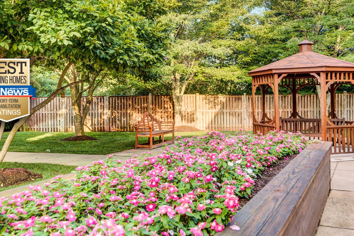 A peaceful outdoor garden area with a wooden gazebo, a wooden bench, a flower bed filled with pink flowers, a paved walkway, and a wooden fence in the background surrounded by green trees.