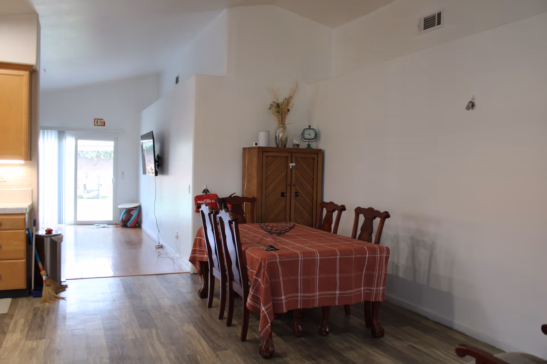 Interior view of a senior care facility dining area with a wooden dining table covered with a red checkered tablecloth and six matching chairs. A wooden cabinet with decorative items on top is against the wall. The room has light-colored walls and wood-patterned flooring. In the background, there is a hallway leading to a glass door with an exit sign above it, and a wall-mounted TV is visible on the left side of the hallway.