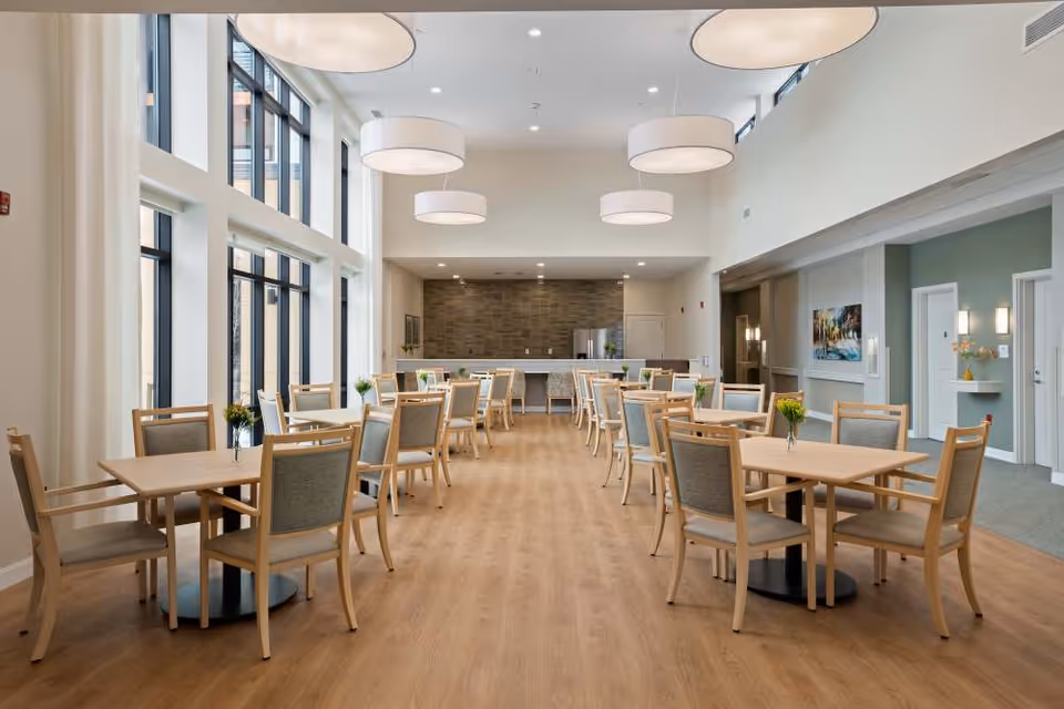 A spacious, well-lit dining room with multiple wooden tables and chairs arranged neatly. Large windows on the left side allow natural light to fill the room. Modern circular ceiling lights hang from the high ceiling. The back wall features a kitchen area with a refrigerator and a counter. Small flower vases are placed on each table.