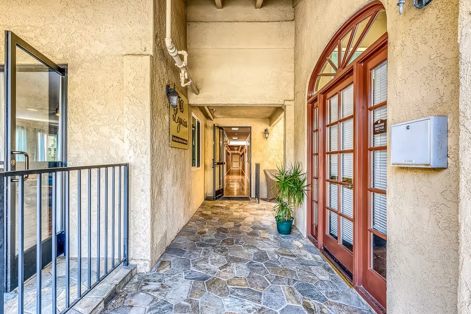 A covered outdoor corridor with stone tile flooring leading to a hallway inside a building. On the right side, there is a set of red-framed glass double doors with a sign that reads 'Executive Director'. A potted plant is placed near the doors. On the left side, there is a black metal railing and an open glass door. The walls are textured and painted beige, with a light fixture and a sign that says 'La Loggia'.