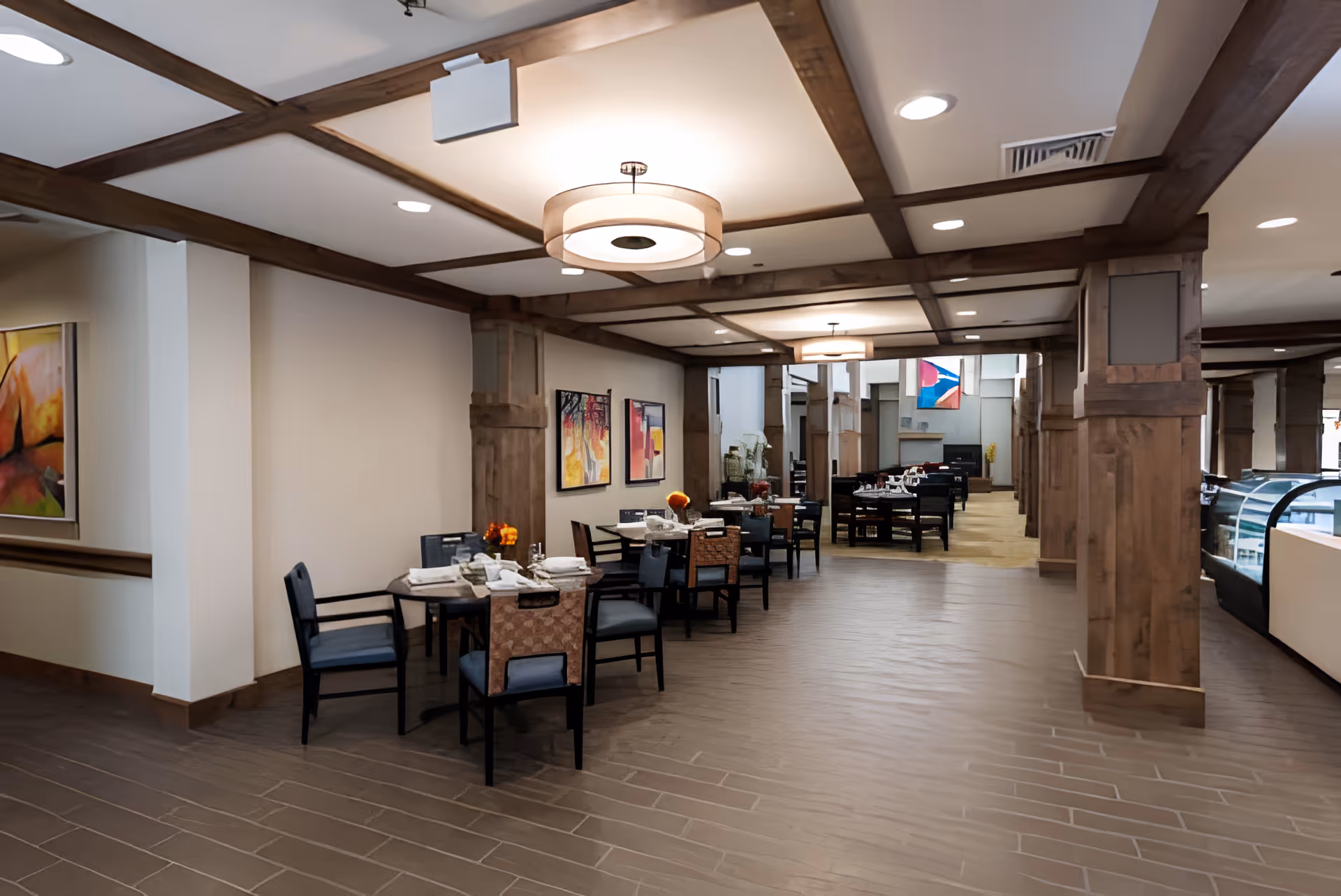 Interior view of a dining area in a senior living facility with multiple tables and chairs arranged neatly. The space features wooden beams on the ceiling, modern light fixtures, and colorful abstract paintings on the walls. Tables are set with napkins and small floral centerpieces, creating a welcoming atmosphere.