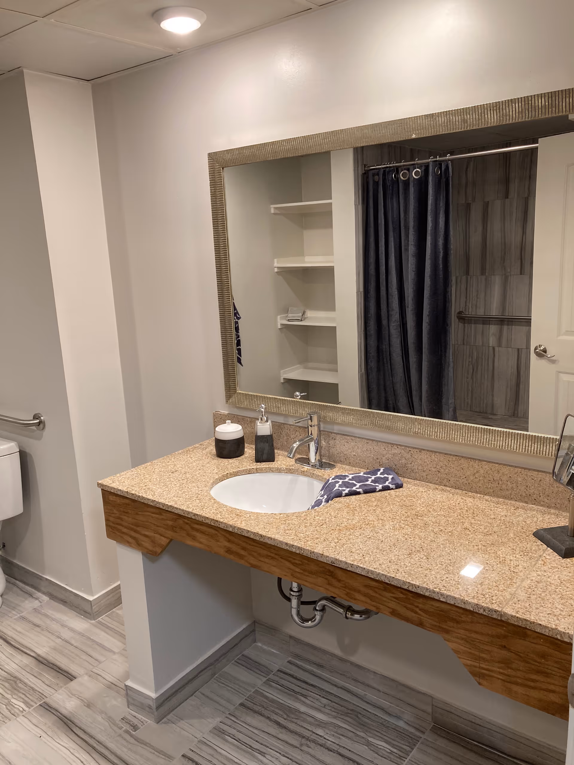 A clean bathroom with a granite countertop sink, a large rectangular mirror, a soap dispenser, a container, and a patterned hand towel. In the background, there is a shower with a dark curtain and tiled walls, a white door, and built-in shelves.