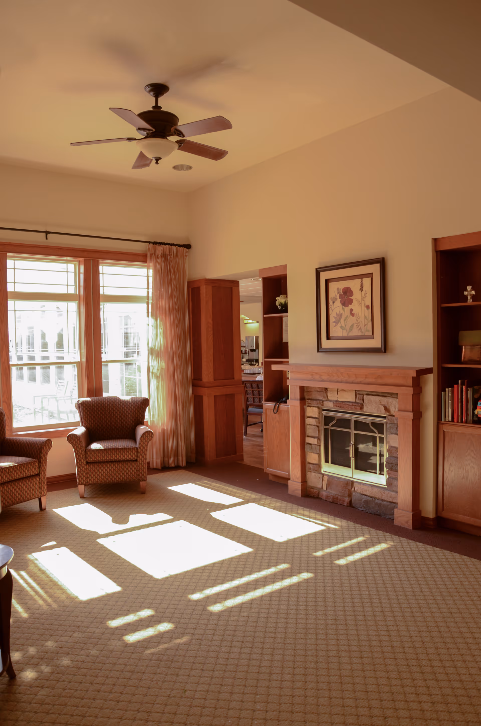 Sunlit living room with armchairs, a fireplace, built-in shelving, and large windows.