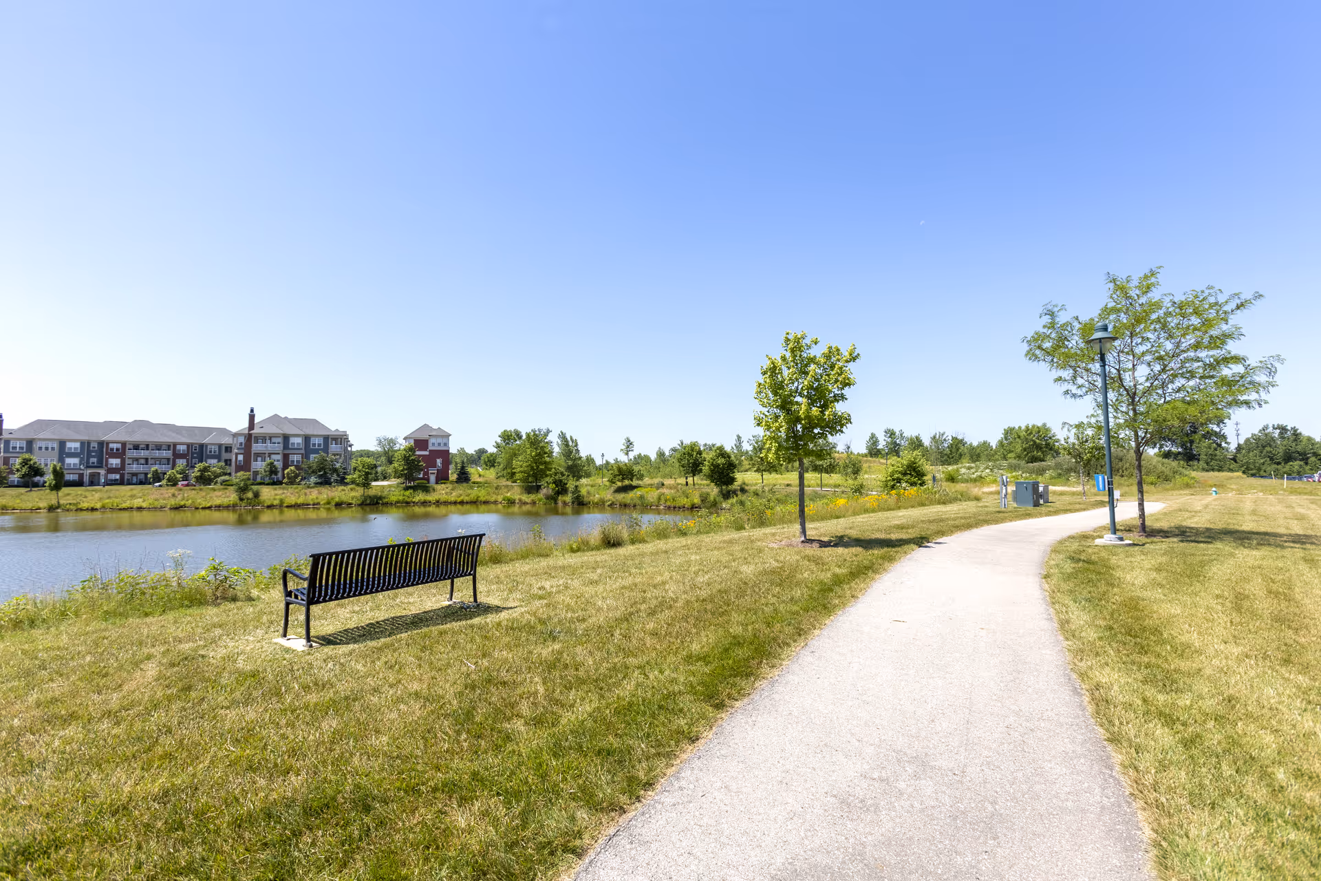 A paved walking path curves through a grassy area with small trees and a black metal bench facing a pond. In the background, there is a multi-story residential building under a clear blue sky.